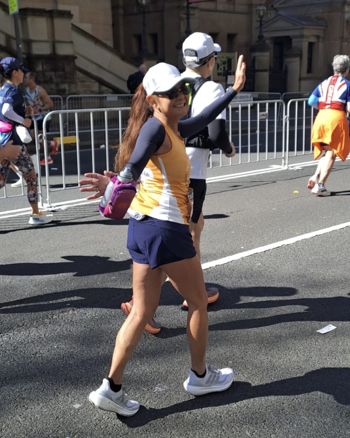 Woman participating in the Sydney Marathon smiles and waves mid-race, wearing a white cap, sunglasses, and an orange running singlet, with runners and spectators in the background.