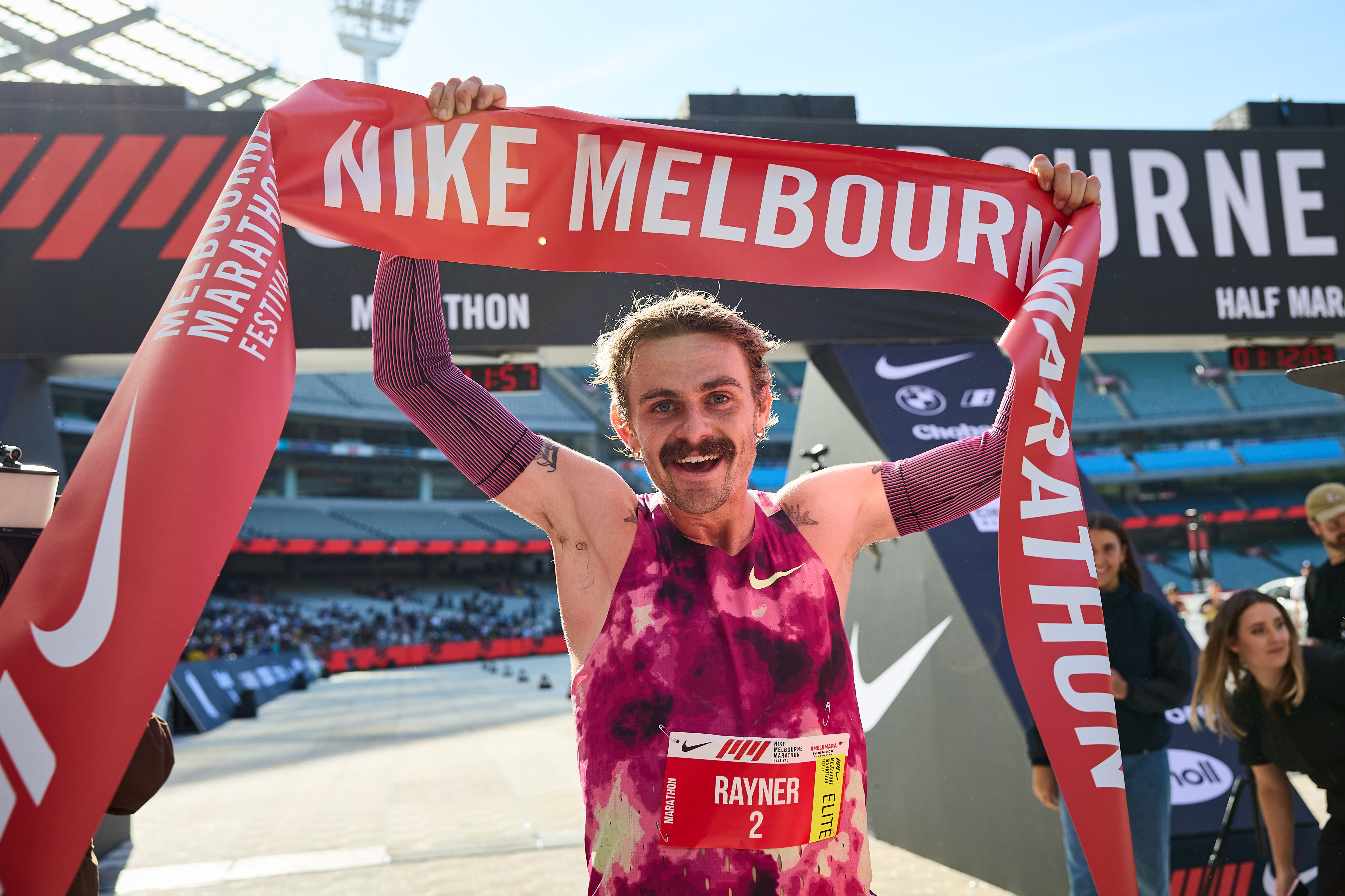 Triumphant moment of a runner in a tie-dye singlet crossing the finish line of the Nike Melbourne Marathon holding a red ‘Nike Melbourne’ ribbon above his head.