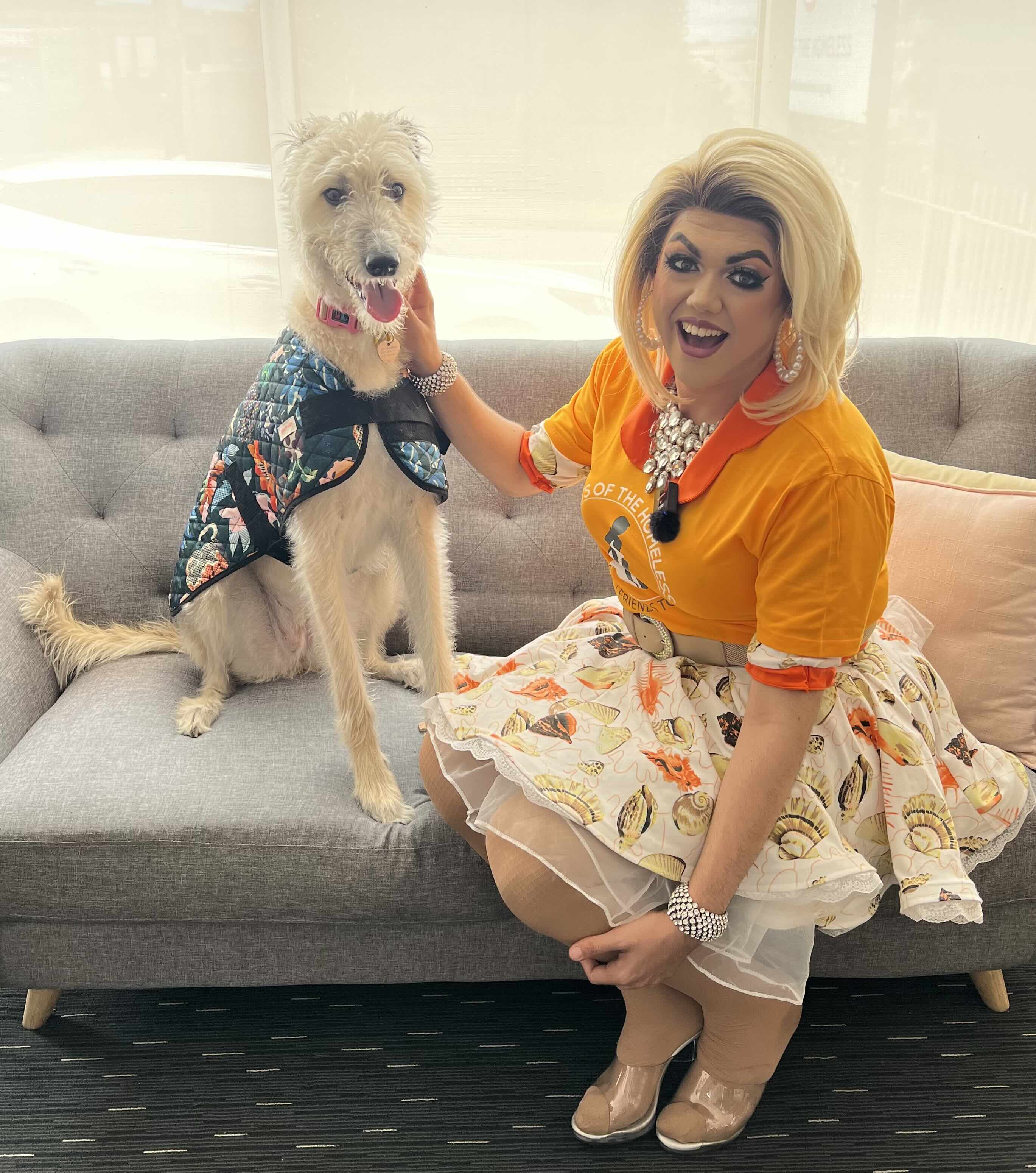 Drag performer in vibrant outfit smiling on a couch with a happy dog for a Pets Of The Homeless Drag Bingo fundraiser event