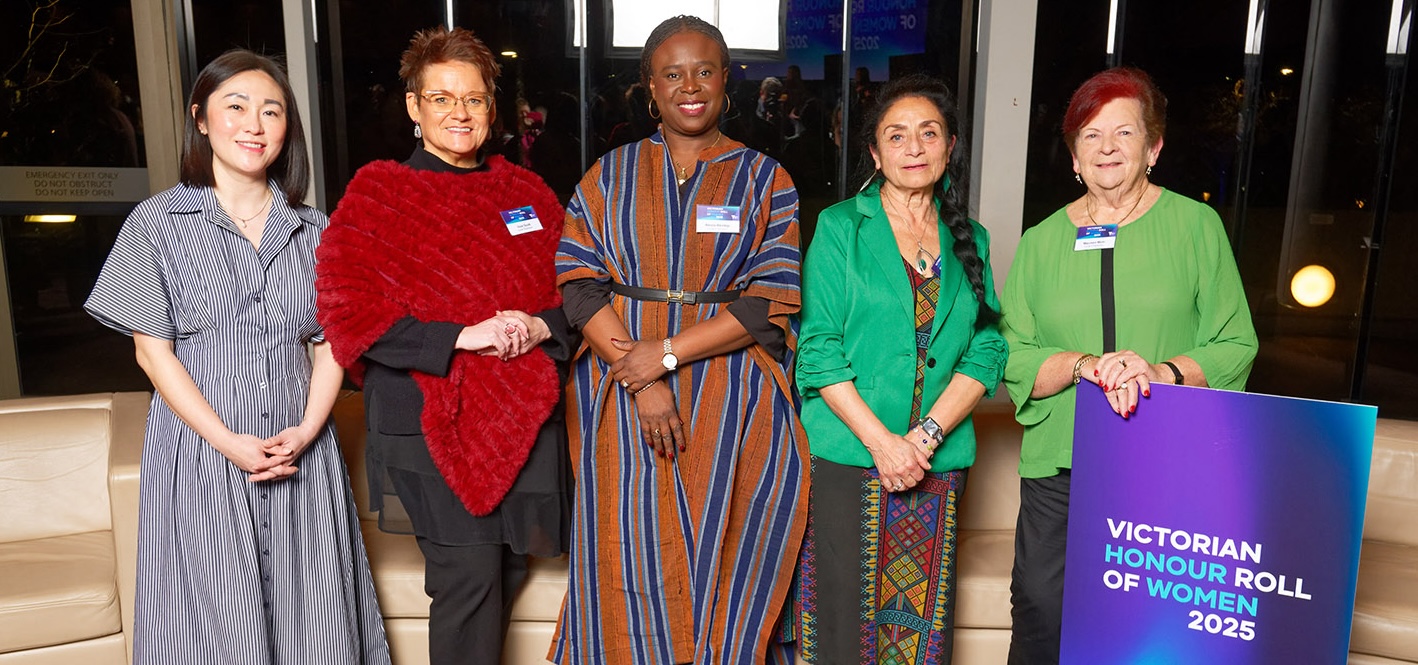 Five women standing proudly in a row, dressed in vibrant outfits, at the Victorian Honour Roll of Women 2025 event. A large sign next to them reads “Victorian Honour Roll of Women 2025”.